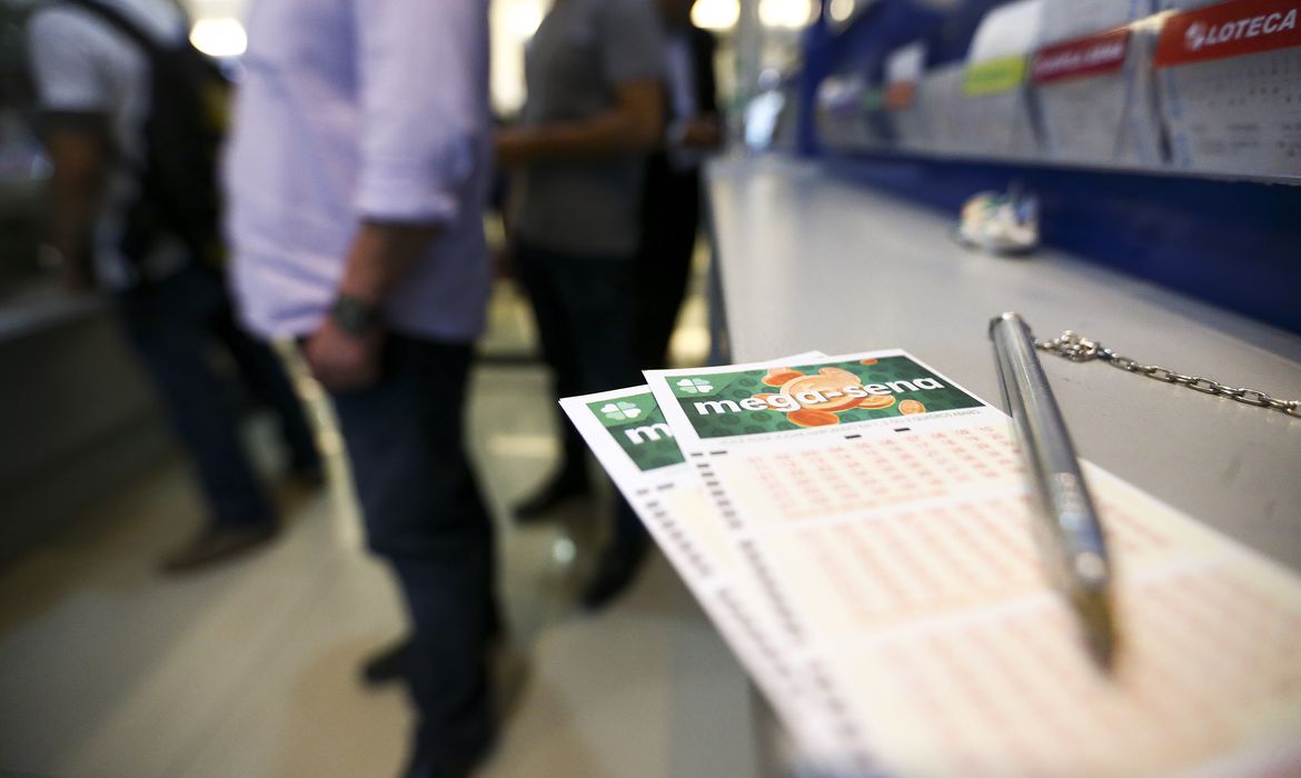 Bilhete de aposta da Mega-Sena em primeiro plano com pessoas na fila de agência lotérica ao fundo