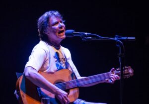 O cantor e compostor Lô Borges tocando violão acústico e cantando em um palco escuro, iluminado por luzes de apresentação. Veste camiseta branca com estampa azul e a palavra "SAUDADE" visível. A imagem transmite emoção e envolvimento artístico durante uma performance musical ao vivo.