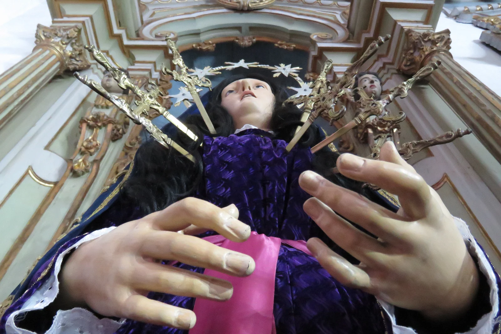 A imagem mostra, em primeiro plano e em ângulo de baixo para cima, uma escultura religiosa de Nossa Senhora das Dores, posicionada em um altar ornamentado de igreja barroca. A Virgem Maria é representada com expressão de sofrimento, olhos voltados para o alto e lábios entreabertos, transmitindo dor e devoção. Sete espadas douradas estão cravadas em seu peito, simbolizando as sete dores de Maria na tradição católica. A imagem veste um manto roxo, com detalhes em tecido que remetem ao período litúrgico da Semana Santa, e uma faixa rosa na altura da cintura. As mãos da escultura estão estendidas à frente, com os dedos delicadamente posicionados, reforçando o gesto de dor e entrega. Ao fundo, o altar apresenta rica ornamentação em estilo barroco, com colunas, entalhes dourados e elementos decorativos que emolduram a imagem. A perspectiva da fotografia valoriza a imponência da escultura e a intensidade emocional da cena, destacando a espiritualidade e a tradição religios