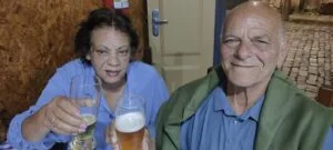 Um casal idoso está sentado lado a lado em um ambiente interno, possivelmente um bar ou pizzaria, brindando com bebidas. À esquerda, a mulher, de cabelos escuros e curtos, veste uma blusa azul e segura um copo de vidro com uma bebida clara. À direita, o homem, de cabelos grisalhos e expressão sorridente, usa camisa azul e tem um casaco verde apoiado sobre os ombros, segurando um copo de cerveja com espuma. Ambos olham em direção à câmera enquanto erguem os copos em um gesto de celebração. Ao fundo, há uma parede com textura rústica e uma porta clara, além de um mural que remete a uma rua de pedra, típico de cidades históricas como Ouro Preto. A cena transmite um clima de alegria, companheirismo e comemoração.