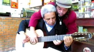 Imagem mostra os músicos Toninho e Jacob durante apresentação intimista em um bar no bairro Barra, em Ouro Preto, em 2014. Os dois aparecem abraçados enquanto tocam o mesmo violão “a quatro mãos”, demonstrando sintonia e cumplicidade musical. Toninho, sentado, segura o instrumento enquanto Jacob acompanha os acordes por trás, com os braços sobre os ombros do parceiro. O ambiente simples do comércio, com balcão, garrafas e objetos ao fundo, reforça o clima espontâneo e afetivo do encontro registrado pelo Jornal Voz Ativa.