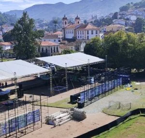 Vídeo mostra a estrutura em montagem para o show do cantor Dilsinho no estacionamento do Centro de Convenções da UFOP, em Ouro Preto. A imagem, captada de um ponto elevado, revela grandes tendas brancas cobrindo parte da área do evento, além de estruturas metálicas, gradis e equipamentos sendo preparados para receber o público. Ao fundo, destaca-se a paisagem característica de Ouro Preto, com casarões coloniais, igrejas históricas e morros cobertos por vegetação. O cenário evidencia os preparativos finais para a realização do evento, com ampla área destinada ao palco e aos setores de circulação dos espectadores.