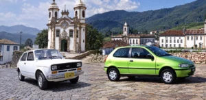 Fiat 147 branco e Fiat Palio verde estacionados sobre calçamento de pedra, tendo ao fundo igrejas, casarões coloniais e montanhas de Ouro Preto, em Minas Gerais.